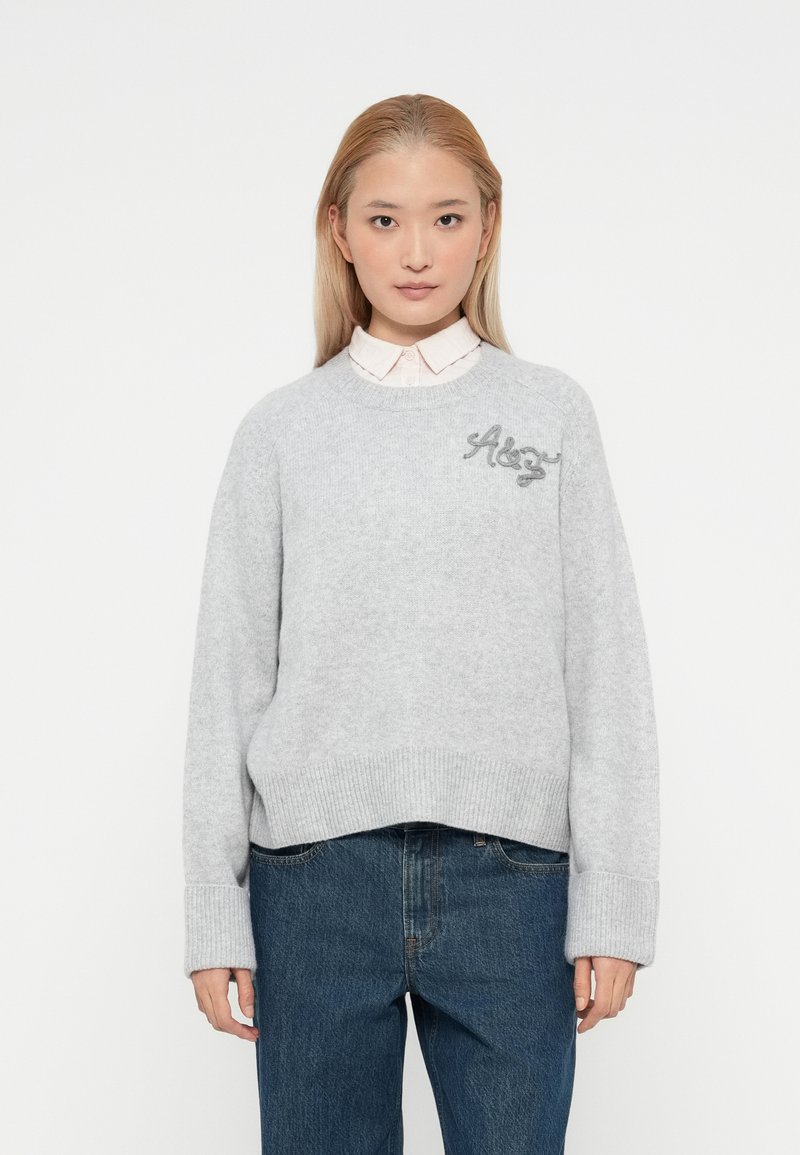 Un pull en maille gris avec un ourlet et des poignets côtelés, présentant un texte brodé sur la poitrine. Porté par-dessus une chemise à col claire.