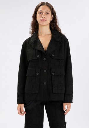 Donna con capelli lunghi e ricci indossa una giacca nera testurizzata con ampi rever, chiusura con bottoni e quattro tasche frontali, abbinata a pantaloni coordinati.