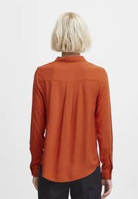 Chemise à manches longues de couleur orange brûlé, confectionnée dans un tissu lisse. Elle présente un col classique, des poignets boutonnés, et de subtiles plissures au dos.