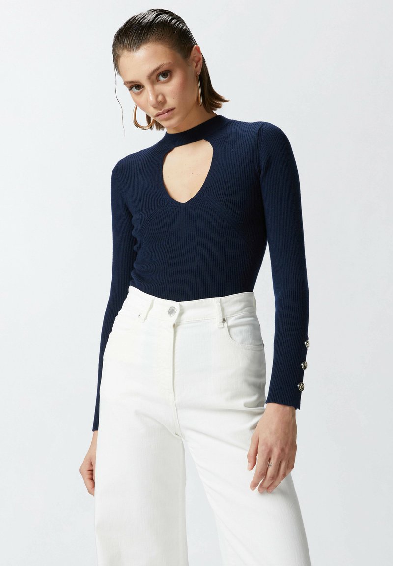 Maglietta a maniche lunghe a coste navy con scollatura a chiave, abbinata a pantaloni bianchi a vita alta con dettagli di bottoni argentati.