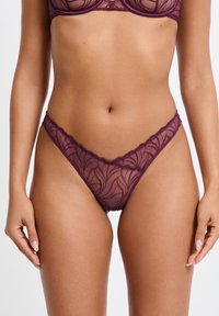String en dentelle violette avec un motif floral, des bords festonnés et une texture semi-transparente, mettant en valeur un design délicat et féminin.