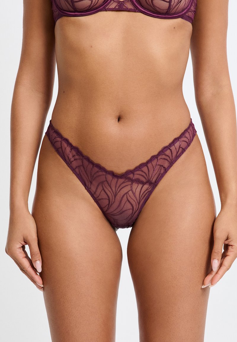 String en dentelle violette avec un motif floral, des bords festonnés et une texture semi-transparente, mettant en valeur un design délicat et féminin.