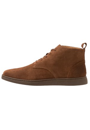 Bottes chelsea en daim marron avec une texture lisse, bout rond, six œillets pour les lacets et une semelle en caoutchouc. Dispose d'une tirette à l'arrière.