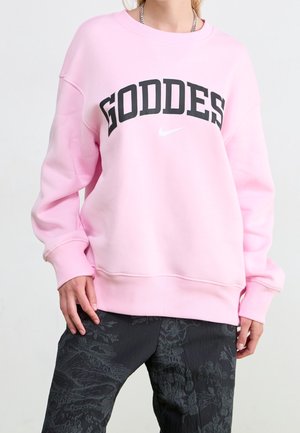 Osoba ubrana w pastelowo różową bluzę z napisem „GODDESS” czarnymi literami i małym białym logo Nike, w połączeniu z czarnymi spodnimi w wzory.