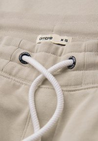 Beige joggers met een trekkoordsluiting en geribbelde tailleband; "ombre" label zichtbaar met maatdetails. Soepele stofstructuur.