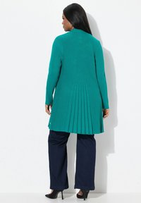 Teal ribbad cardigan med veckad ryggdetalj, långa ärmar, ihop med mörkblå vida byxor och svarta klackar.