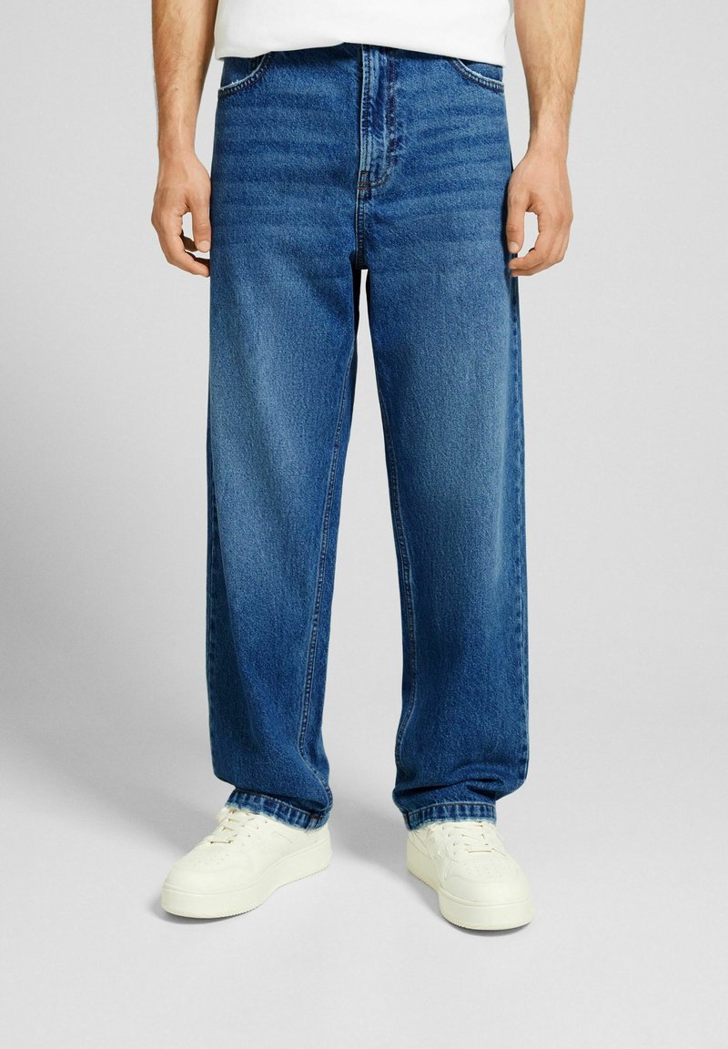 Jeans a gamba larga in denim blu con vestibilità a vita media, dotati di tasche frontali e leggeri dettagli sbiaditi, abbinati a sneakers bianche.