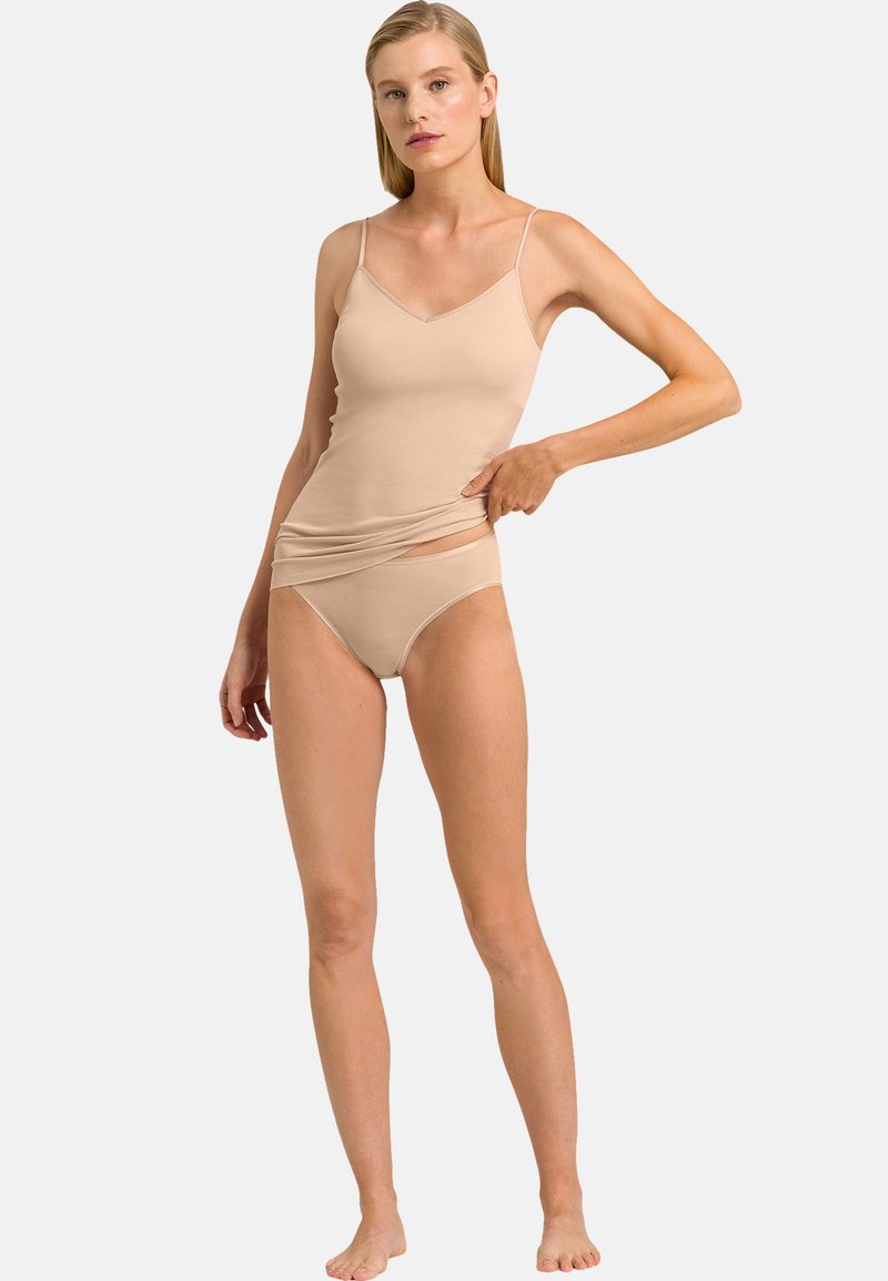 Femme portant un caraco beige et des sous-vêtements assortis, tirant légèrement sur le caraco pour révéler la ceinture, debout pieds nus sur un fond clair.