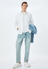 Camisa blanca de botones, jeans azul claro, chaqueta de denim azul claro colgada del brazo y zapatillas blancas sobre un fondo liso.