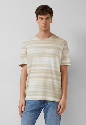 Jonge man die een beige-wit horizontaal gestreept T-shirt met korte mouwen en blauwe jeans draagt, staande tegen een effen lichtgrijze achtergrond.