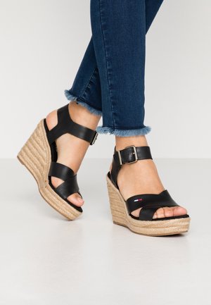 Sorte kilehælssandaler med flettet jutedetalje, der har brede stropper og en ankelspænde. Kombineret med mørkeblå jeans med frynset kant.