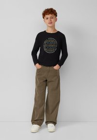 Zwarte langärmlige shirt met een cirkelvormige "SOUND" graphic, gecombineerd met lichtbruine wijde broek en witte sneakers.