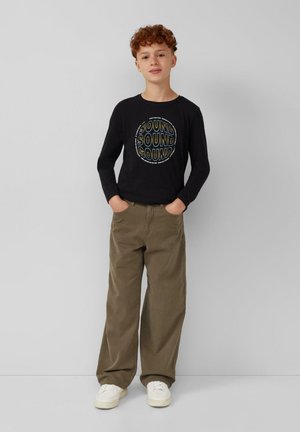Zwarte langärmlige shirt met een cirkelvormige "SOUND" graphic, gecombineerd met lichtbruine wijde broek en witte sneakers.
