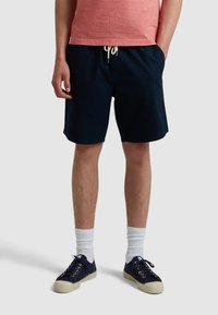 Herren Shorts in Marineblau mit elastischem Bund und Kordelzug. Getragen mit einem roten Oberteil, weißen Socken und marineblauen Schuhen mit hellen Sohlen.