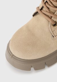 Botte en suede beige avec une pointe ronde, surface texturée et semelle en caoutchouc épaisse dotée de profondes rainures pour une meilleure adhérence. Lacets marron sécurisant le dessus.