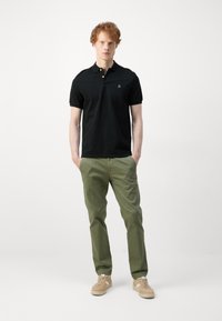 Camisa polo negra de manga corta y cuello, combinada con pantalones verde oliva. Tejido suave con un pequeño logo en el pecho. Calzado casual.