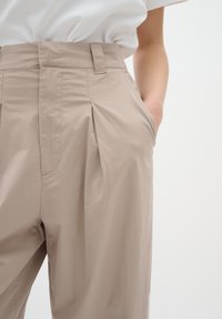 Béžové kalhoty s předními sklady, bočními kapsami a hladkou texturou látky. Pás má poutka na opasek pro nastavení.