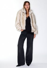 Giacca in pelliccia ecologica color crema, caratterizzata da un colletto alto e ampie maniche, indossata sopra un top nero in pizzo e abbinata a jeans neri a gamba larga.