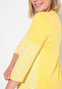 Gul topp med trekvartsärmar; en ärm har vita ränder. Slät textur, med en figurnära design och rund halsringning.
