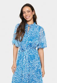Robe légère, bleue, à motifs avec des manches courtes et flottantes, un col à nouer et une taille cintrée. Le tissu semble transparent et fluide.