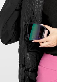 Volcom skibril met een zwart frame en een meerkleurige, spiegelende lens, gehouden in een ritssluiting van een zwarte jaszak.