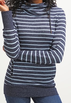 Kobieta ubrana w granatowy i jasnoniebieski sweter w paski z długim rękawem, z kołnierzem kapturowym i ściągaczem, zestawiony z niebieskimi dżinsami.