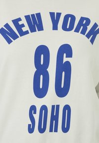Camiseta de algodón blanca con gran texto azul: "NUEVA YORK" arqueado sobre "86 SOHO." Diseño sencillo con un estilo casual.