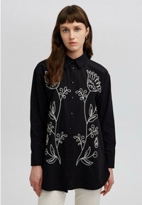 Black shirt with long sleeves, button closure, and white floral embroidery on the front. Soft fabric with a relaxed fit.