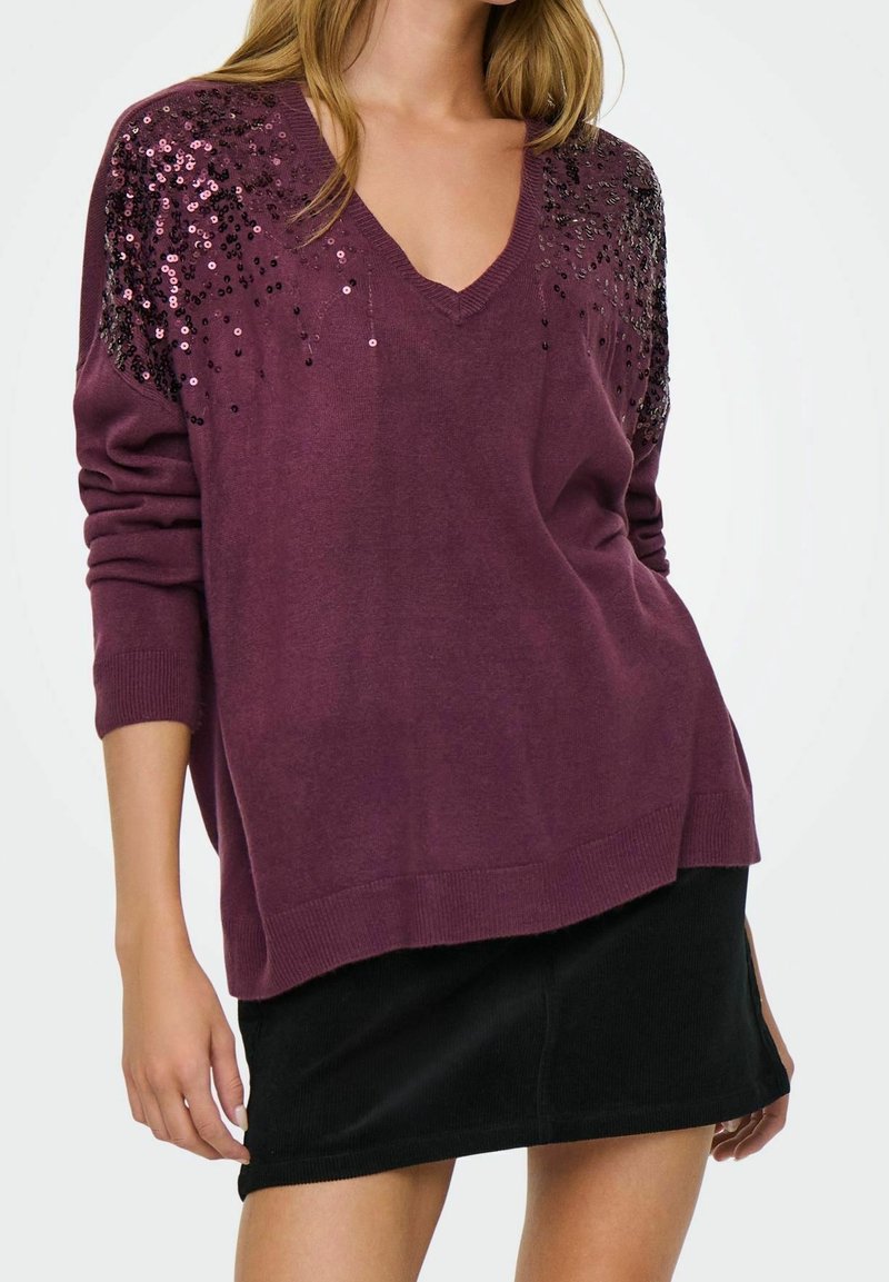 Femme portant un pull violet ample à col en V avec des sequins sur les épaules et une jupe courte noire, debout devant un fond uni.