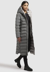 Grå polstret lang frakke med en stor hætte, der har en frontlynlås, frontlommer og en glat inderside. Båret over sort tøj og støvler.