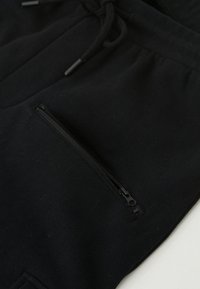 Čierne joggingové nohavice vyrobené z mäkkej látky, s predným vreckom na zips na stehne a nastaviteľnými šnúrkami v oblasti pása.