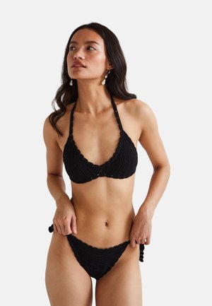 Femme portant un bikini noir en crochet avec un haut halter et un bas avec liens sur les côtés, regardant légèrement vers sa gauche sur un fond uni.