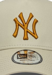 Μπεζ καπέλο με εμφανές κίτρινο κεντητό λογότυπο "NY". Περιλαμβάνει κυκλικό ετικέτα με τη φράση "New Era" και "Ρυθμιζόμενο". Ομαλή υφή υφάσματος.