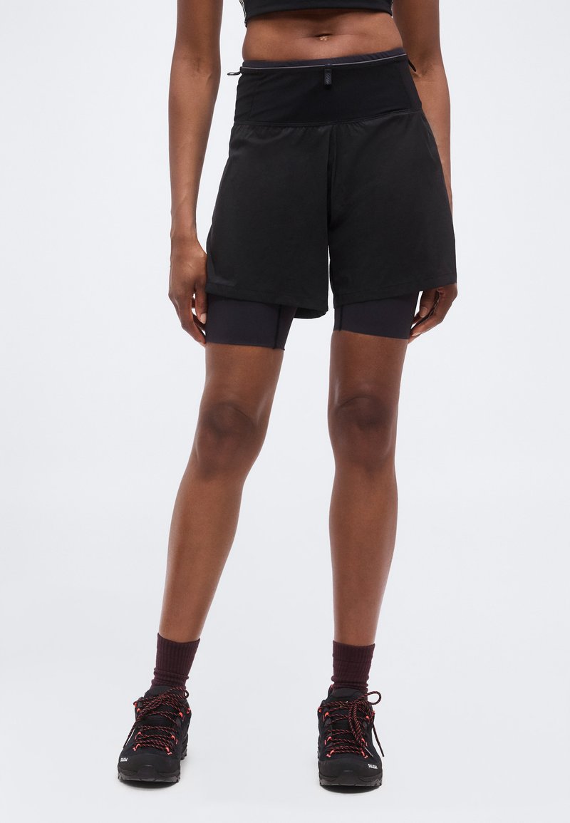 Αθλητής που φοράει μαύρα στρωμένα σορτς για τρέξιμο με εσωτερικά συμπιεστικά σορτς, μαύρα και πορτοκαλί παπούτσια για τρέξιμο σε μονοπάτι, στέκεται μπροστά σε απλό φόντο.