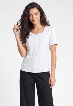 Vrouw met lang, golvend haar draagt een wit blouse met korte mouwen, een geplooide halslijn en een zwarte broek, staat en lacht tegen een egale achtergrond.