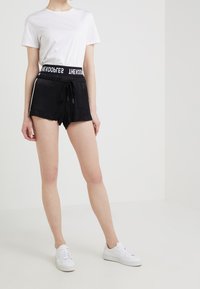 Persona che indossa una maglietta bianca a maniche corte, pantaloni corti neri con coulisse e fascia con logo, e sneakers bianche, in piedi davanti a uno sfondo bianco.