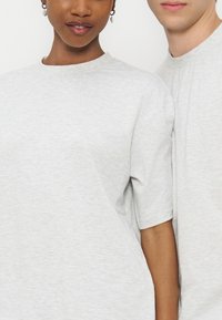 Deux personnes portant des t-shirts gris clair à col rond, visibles des épaules jusqu'au milieu du torse, debout proches l'une de l'autre.