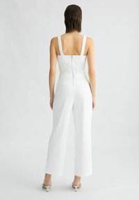 Salopette blanche avec pantalon à jambes larges, un buste ajusté avec une fermeture éclair au dos, et de larges bretelles, avec des détails de tissu texturé le long de la taille.