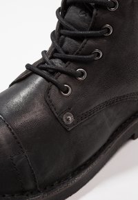 Černá kožená kotníková bota s texturovaným povrchem, černými šňůrkami, kovovými očky a vyztuženou špičkou. Obsahuje šitý podpatek.