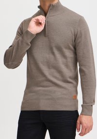 Maglione marrone con zip e colletto e polsini a costine. Texture liscia, design aderente, con un'etichetta in pelle sul lato inferiore.