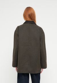 Σκούρο πράσινο παλτό με γιακά και μακριά μανίκια, με μινιμαλιστικό σχεδιασμό. Το ύφασμα έχει λεία υφή και ευθεία γραμμή.