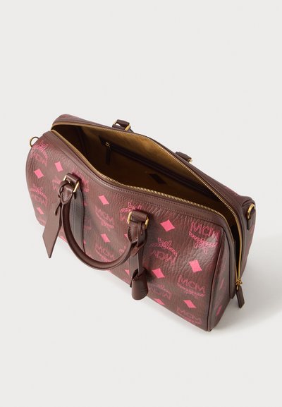 Καφέ δερμάτινη τσάντα duffel με ροζ λογότυπα MCM, χρυστό φερμουάρ μερικώς ανοιχτό, διπλές λαβές και εσωτερική τσέπη με φερμουάρ ορατή στο εσωτερικό.