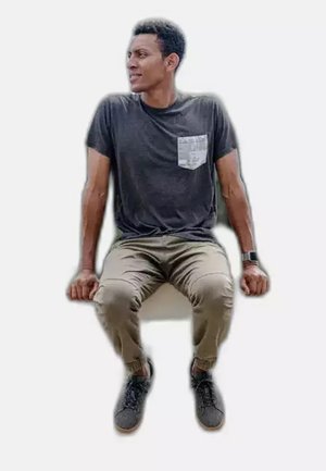 Young man sitting with hands gripping edge, wearing black t-shirt with patterned pocket, beige pants, and dark sneakers, looking left.