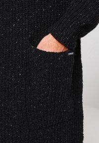 Černý pletený svetr s ribbed texturou, s kapsou na přední straně a malou logo etiketu. Příze má vícero barevné skvrny.