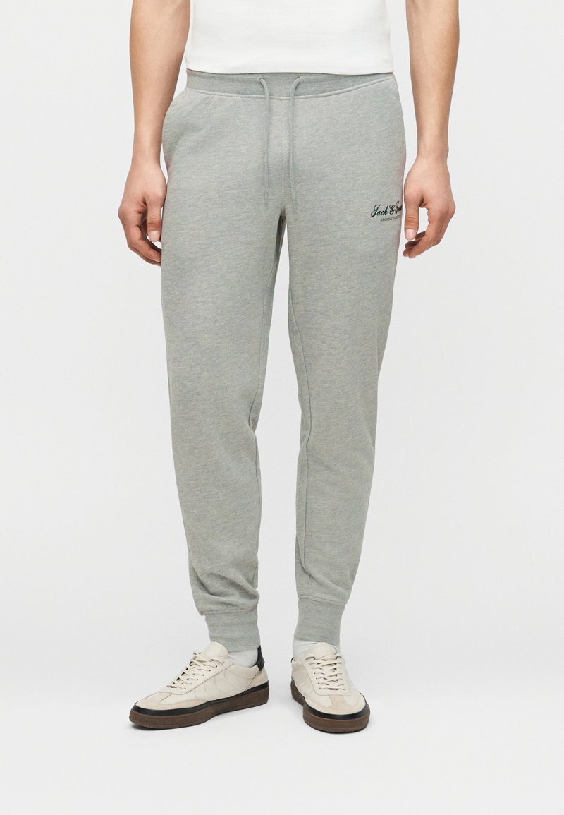 Άνδρας που φοράει ανοιχτό γκρι παντελόνι φόρμας με σούρα στη μέση και μανσέτες στους αστραγάλους, συνδυασμένο με λευκά αθλητικά παπούτσια και λευκές κάλτσες.