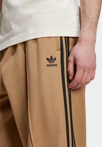 Pantalon de sport marron en tissu texturé, avec des rayures latérales noires et un logo Adidas brodé sur la cuisse gauche.