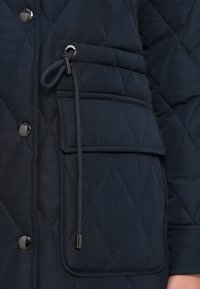 Gros plan sur une veste matelassée marine montrant des boutons-pressions métalliques, un cordon de serrage réglable avec des embouts en métal, et une grande poche à rabat sur la manche.