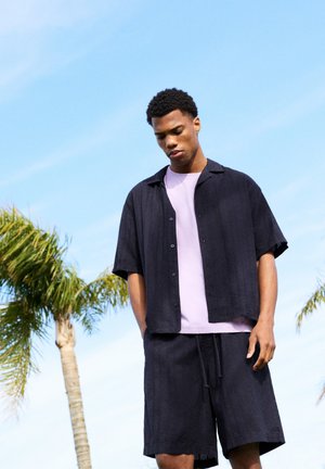 Jeune homme en chemise noire à boutons ouverts et short se tient à l'extérieur, les mains dans les poches, face à un ciel bleu et des palmiers.
