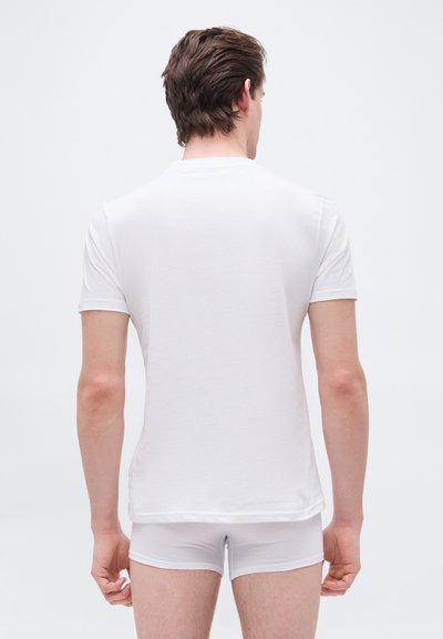 Άνδρας με την πλάτη γυρισμένη, φοράει απλό λευκό κοντομάνικο T-shirt και λευκά εσώρουχα μπόξερ, σε ουδέτερο φόντο.