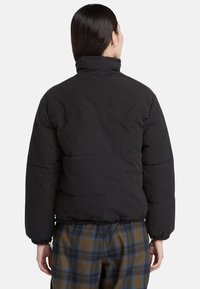 Giacca a vento nera con colletto alto, design imbottito e polsini elasticizzati. Indossata sopra pantaloni a quadri con motivi marroni e blu.